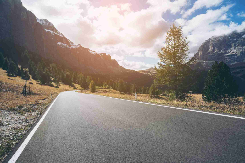 A winding road curves through a sunlit mountain valley, bordered by trees under a bright sky. The peaceful landscape conveys serenity and freedom.